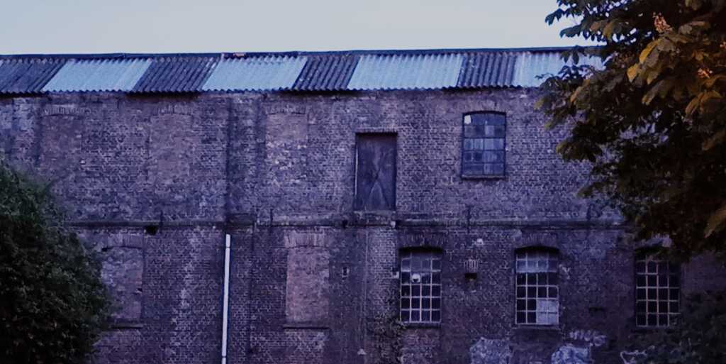 A photo of an abandoned warehouse taken early in the morning. The dark brick building has several sealed doors, some of which are inaccessibly high up. There are large framed windows all across the wall.