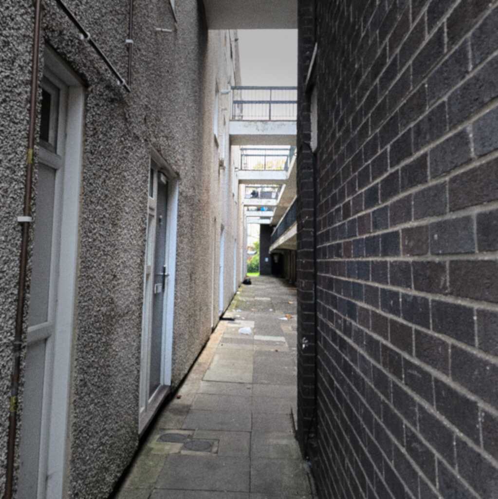 A brightly lit view into a small, narrow residential block. Repeated overhead walkways can be seen stretching into the distance. On the left is a concrete light grey block with doors. On the right is a dark grey brick wall.